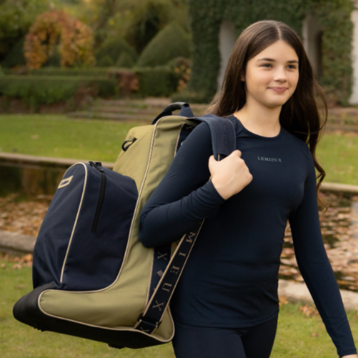 LeMieux AW26 Boot & Hat Bag Varient Gooseberry. A young woman walking outdoors in a garden while carrying a large navy and olive green hat and boot bag over her shoulder. She is wearing a navy long-sleeve athletic shirt with the brand name 'LeMieux' visible on the chest.