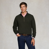 Holland Cooper Quarter Zip Sweat (Men's) Variant Racing Green. A man wearing a dark olive green quarter-zip sweater over a pink collared shirt, paired with blue jeans and a brown belt, standing against a textured light gray background.