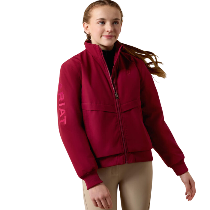 Ariat Youth 2.0 Insulated Stable Jacket Variant Beet Red. A girl wearing a burgundy zip-up jacket with 'ARIAT' printed in pink on the sleeve, paired with beige riding pants, against a solid white background.