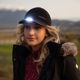 LeMieux AW26 Torch Cap Variant Colour Black. A model with blond long hair wearing a black baseball cap with a torch and black winter jacket on faded countryside background. 