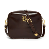 Holland Cooper Country Camera Bag Variant Chesnut Leather. A dark brown glossy leather camera bag with a gold-tone decorative monogram clasp and a matching brown leather shoulder strap, shown on a plain white background.
