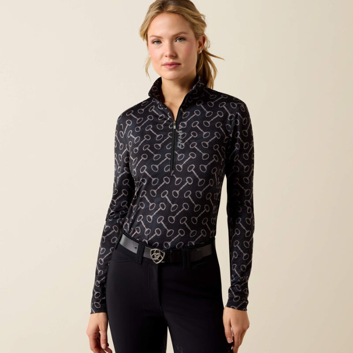 Ariat Ladies Lunaveil 1/2 Zip Long Sleeve Base Layer Variant Black French Link Print. A woman modeling a long-sleeve half-zip performance top with a black base and a white equestrian-style pattern, paired with black riding pants against a solid light beige background.