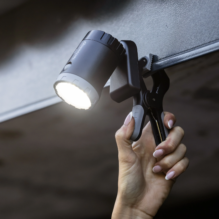 A black clip-on LED work light with a bright white illuminated bulb, held by a hand under a dark metal surface.