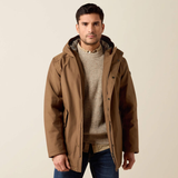 A man wearing a brown hooded parka jacket over a beige knit sweater and blue jeans, standing against a plain light beige studio background.