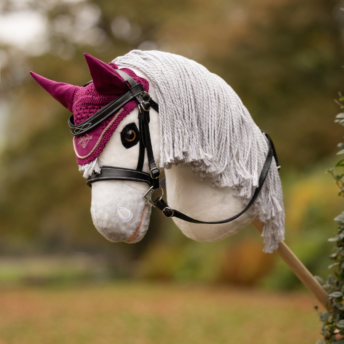LeMieux AW26 Hobby Horse Fly Hood Variant Colour Blackberry. A plush toy horse head on a stick, white colour with wool briaded mane and a blackberry coloiur fly hood and bridle on a faded garden background.
