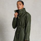 Holland Cooper Chartwell Rain Parka Variant Hunter Green. Lady wearing a green waterproof jacket with gold button detailing against a grey background.