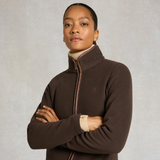 Holland Cooper Country Fleece Jacket Variant Chocolate. A woman wearing a dark brown zip-up fleece jacket with a high collar and a cream ribbed turtleneck underneath, standing with arms crossed against a textured light gray background.