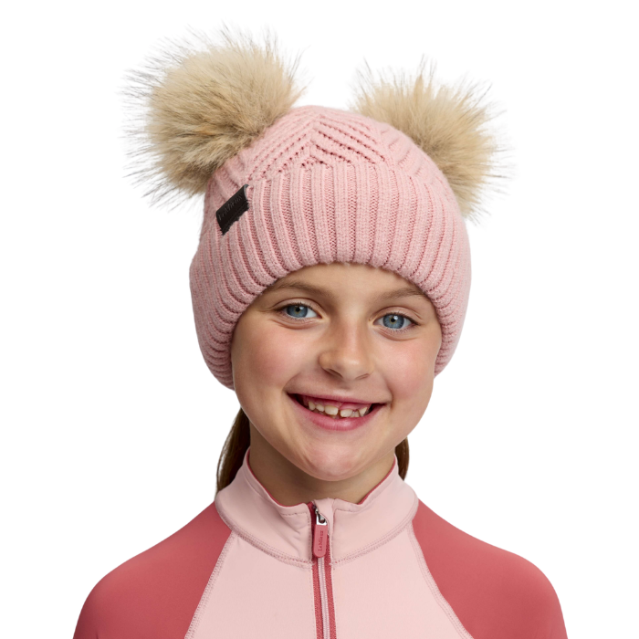 LeMieux AW26 Mini Double Pom Beanie Variant Petal. A smiling young girl wearing a light pink ribbed knit beanie with two large beige faux-fur pom-poms and a small black logo tab, paired with a pink and coral zip-up top against a plain white background.