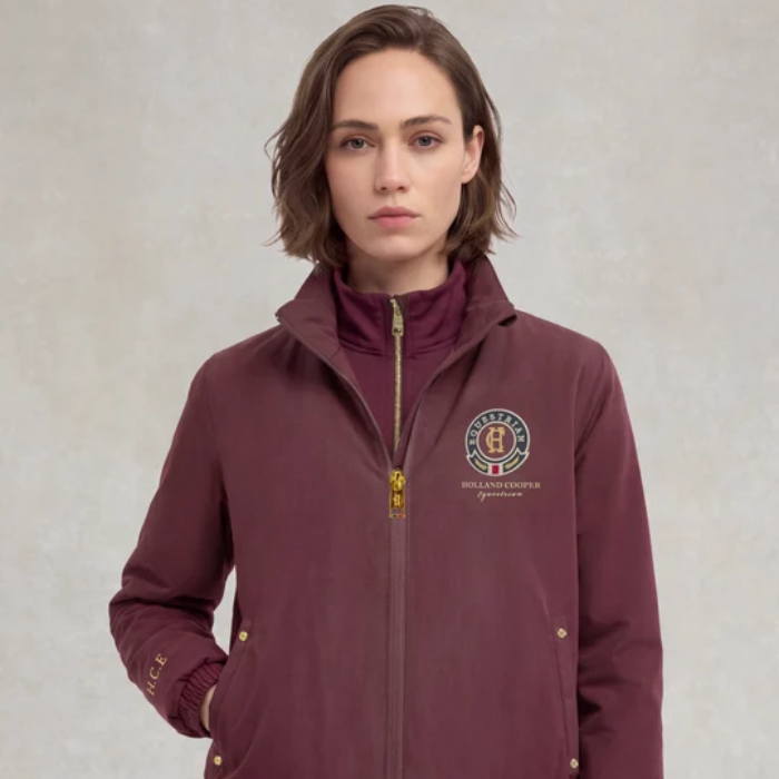 A woman wearing a maroon zip-up jacket with a gold zipper and a circular embroidered crest on the left chest, photographed against a neutral textured gray background.