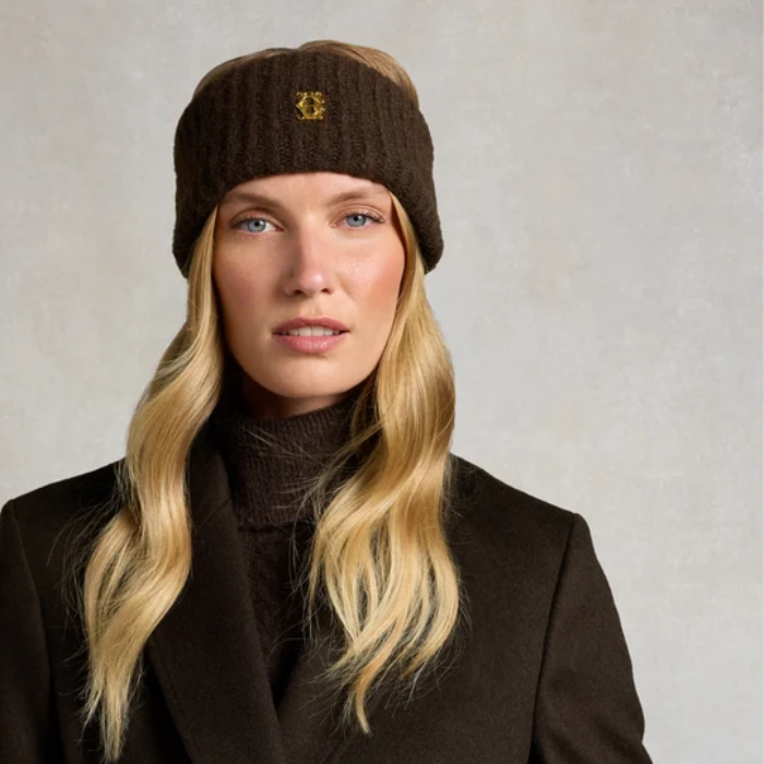 A blonde woman wearing a dark brown ribbed knit beanie with a small gold embroidered crest logo, paired with a dark brown turtleneck sweater and dark coat, set against a neutral textured gray background.