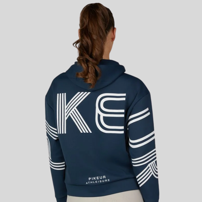 Pikeur AW26 Athleisure Zip Hoody Variant Pine Green. A woman viewed from the back wearing a navy blue hoodie with large white stylized 'PIKEUR' lettering and white stripe graphics on the back and sleeves, plus 'PIKEUR ATHLEISURE' near the lower back, against a light gray studio background.