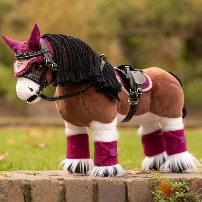 LeMieux AW26 Toy Pony Bandages Variant Colour Blackberry. A plush toy pony wearing an equestrian costume including tack, pad and bandages on faded garden background. 