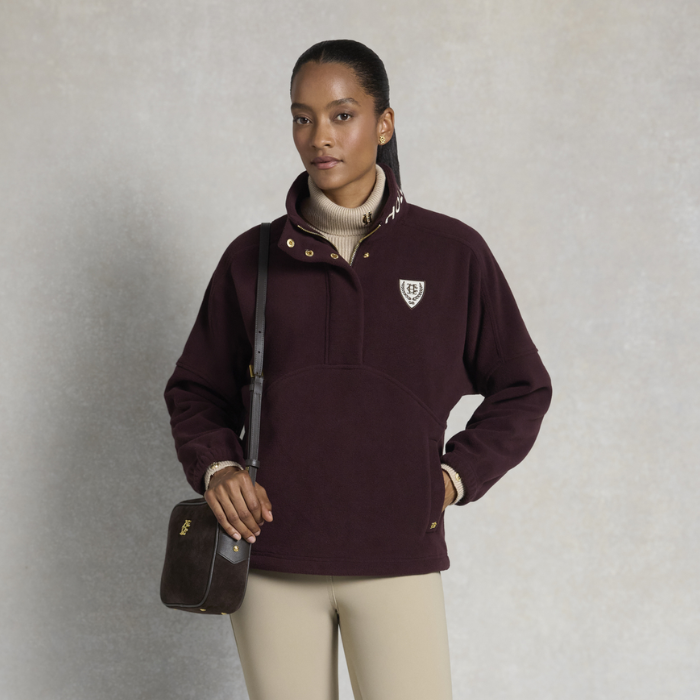 Holland Cooper Cranborne Over Head Fleece Variant Mulberry. A woman modeling a dark burgundy quarter-zip fleece pullover with a small white shield logo on the left chest, paired with beige pants and a dark brown crossbody bag, set against a textured gray studio background.
