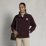 Holland Cooper Cranborne Over Head Fleece Variant Mulberry. A woman modeling a dark burgundy quarter-zip fleece pullover with a small white shield logo on the left chest, paired with beige pants and a dark brown crossbody bag, set against a textured gray studio background.