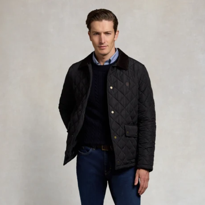 A man wearing a navy quilted jacket with a brown corduroy collar, layered over a dark sweater and blue jeans, standing against a neutral textured background.