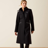A woman modeling a long black hooded coat with reflective silver trim and a small shield logo on the chest, photographed against a plain beige studio background.