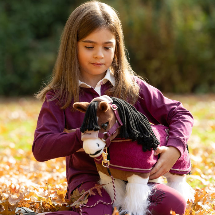 LeMieux AW26 Toy Pony Rug Variant Colour Blackberry. A young person sat on grass with a plush toy pony on their lap. colour theme of outfits in blackberry. on faded garden background