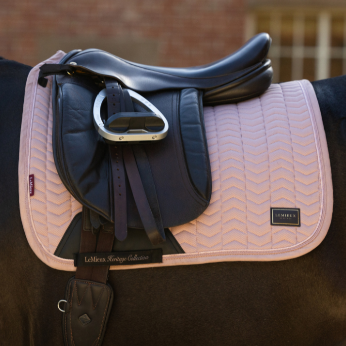 A pink and beige chevron-patterned LeMieux saddle pad with a black leather equestrian saddle and silver stirrup, shown on a horse in front of a blurred brick wall.