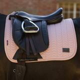 A pink and beige chevron-patterned LeMieux saddle pad with a black leather equestrian saddle and silver stirrup, shown on a horse in front of a blurred brick wall.