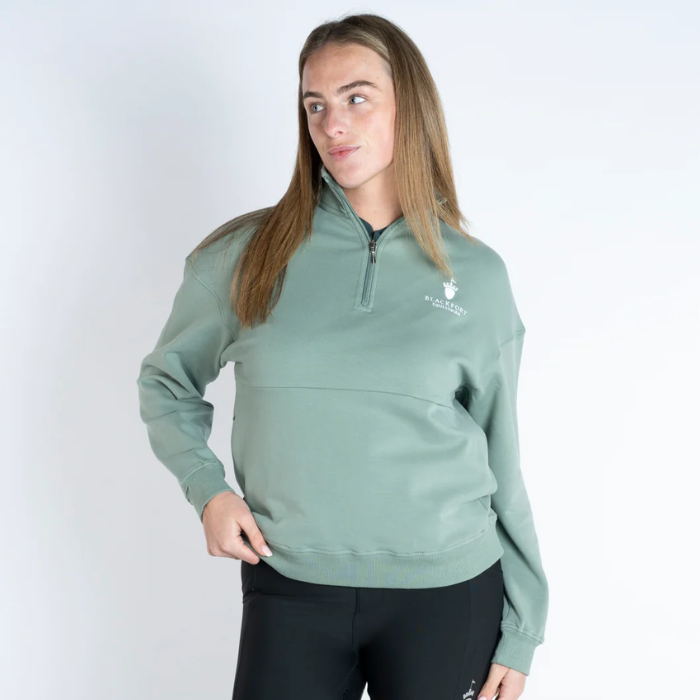 Blackfort Equestrian Ella Quarter Zip Variant Sage. A woman modeling a sage green quarter-zip pullover sweatshirt with a small white embroidered logo on the left chest, paired with black leggings, against a plain white studio background.
