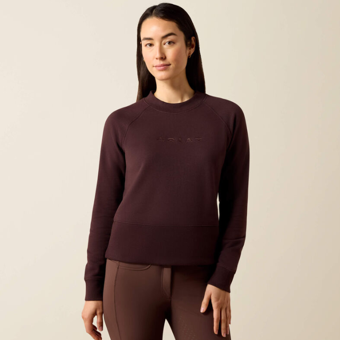 A woman modeling a dark plum crewneck sweatshirt with tonal embroidered branding on the chest, paired with matching leggings, against a solid light beige background.