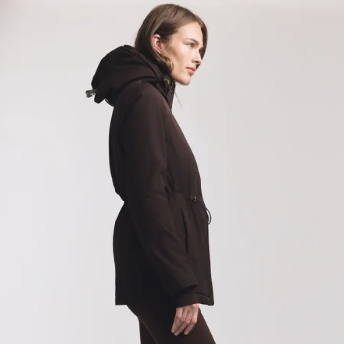 Lady wearing a Brown coat sinched in at the waist against a grey background. 