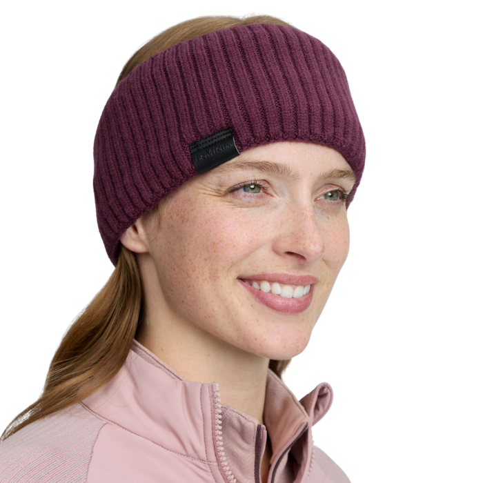 LeMieux AW26 Clara Rib Headband Variant Bramble. A woman wearing a ribbed knit athletic headband in deep plum purple. The headband has a small black fabric brand tag on the side.