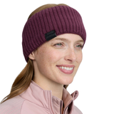 LeMieux AW26 Clara Rib Headband Variant Bramble. A woman wearing a ribbed knit athletic headband in deep plum purple. The headband has a small black fabric brand tag on the side.
