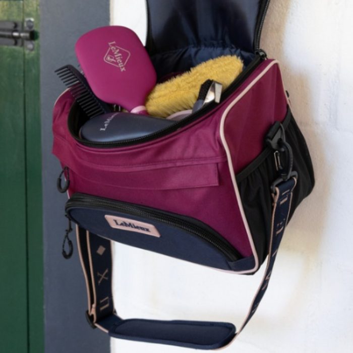 LeMieux AW26 ProKit Lite Grooming Bag Varient Blackberry. A burgundy and navy blue LeMieux bag with an open top, shown hanging on a white wall next to a green door. The bag contains beauty accessories including a pink brush, a yellow brush, and a black comb.