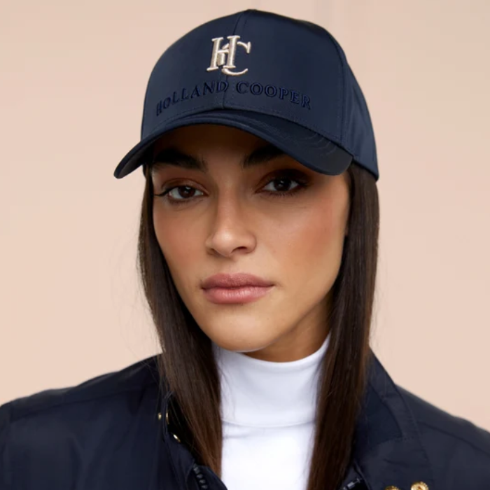 Lady wearing a Navy cap with HC embroided logo against a pink background. 