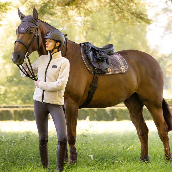 A woman in equestrian attire stands beside a brown horse outdoors in a grassy field. The horse is outfitted with a dark saddle, saddle pad, bridle, and fly veil, while the rider wears a cream fleece jacket, dark helmet, dark breeches, and tall boots.