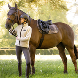 A woman in equestrian attire stands beside a brown horse outdoors in a grassy field. The horse is outfitted with a dark saddle, saddle pad, bridle, and fly veil, while the rider wears a cream fleece jacket, dark helmet, dark breeches, and tall boots.