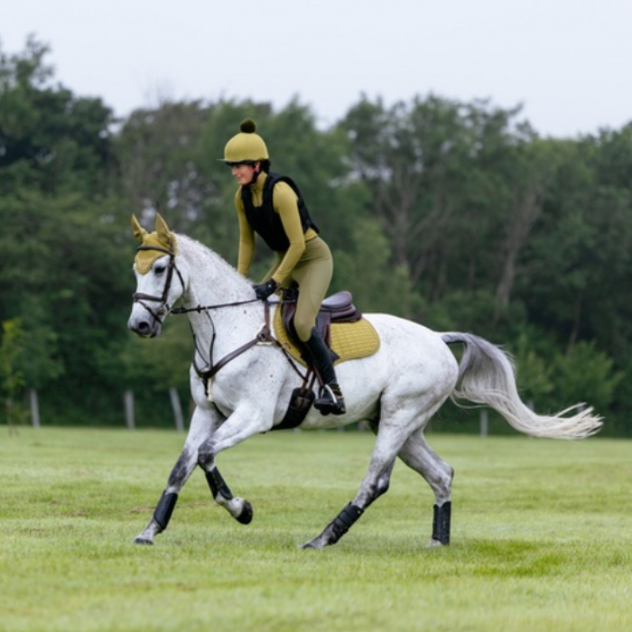 LeMieux AW26 Suede Close Contact Square Varient Gooseberry. A person riding a white horse across a grassy field, wearing a black and olive riding outfit with a matching helmet. The horse has a white and gray coat with black leg wraps, and the background shows trees under an overcast sky.