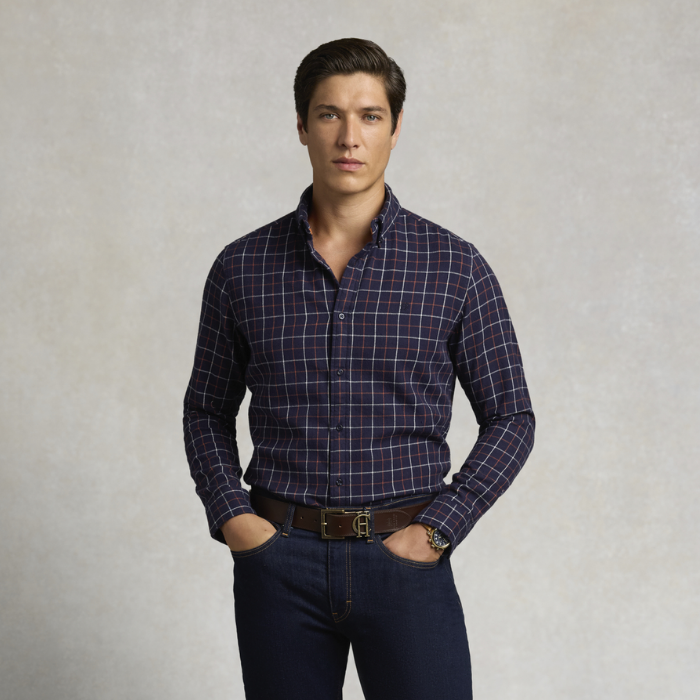 Holland Cooper Men's Classic Fit Brushed Cotton Shirt Variant Copper Navy Check. A man wearing a navy blue long-sleeve plaid button-down shirt with thin red and white lines, paired with dark blue jeans and a brown belt, standing against a textured light gray background.