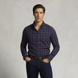 Holland Cooper Men's Classic Fit Brushed Cotton Shirt Variant Copper Navy Check. A man wearing a navy blue long-sleeve plaid button-down shirt with thin red and white lines, paired with dark blue jeans and a brown belt, standing against a textured light gray background.