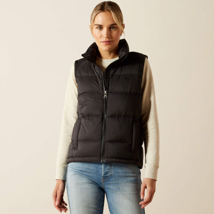 Ariat Women's Ultrapuff Down Vest Variant Black. A woman wearing a black sleeveless puffer vest over a cream long-sleeve top and blue jeans, standing against a plain light beige studio background.