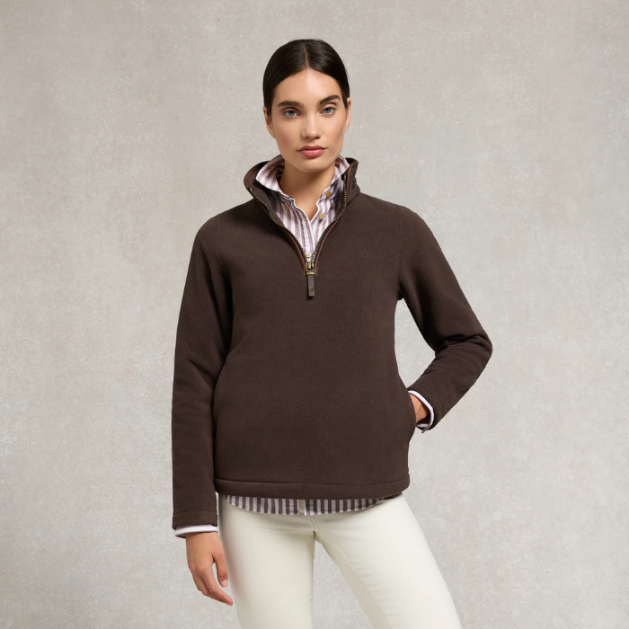 Holland Cooper Ladies Country Fleece Quarter Zip Variant Chocolate. A woman modeling a dark brown quarter-zip fleece pullover sweater, layered over a striped collared shirt and paired with white pants, set against a textured light gray studio background.