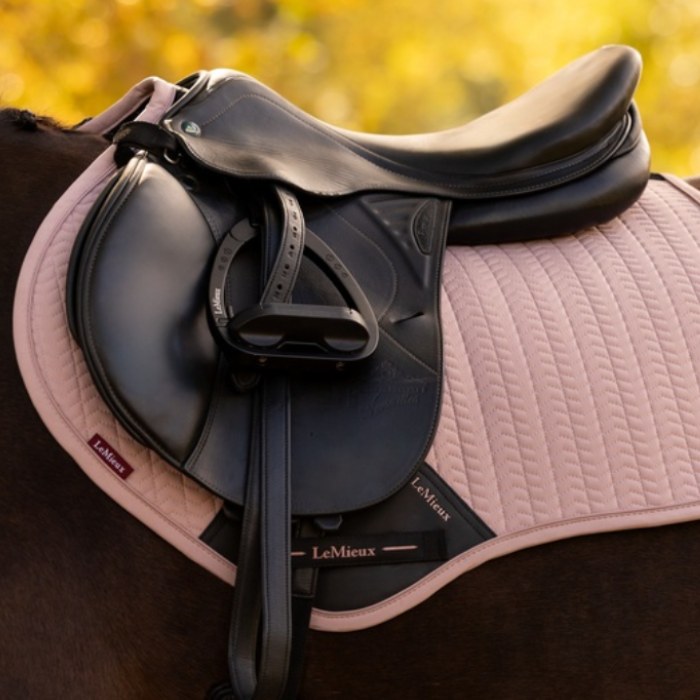 LeMieux AW26 Essence Close Contact Square Varient Wild Rose. A close-up of an equestrian saddle setup on a horse, featuring a black leather saddle and black stirrups mounted on a light pink quilted saddle pad. The LeMieux brand name is visible on the pad and saddle strap, with a warm, blurred golden background.