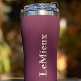 LeMieux AW26 Travel Cup Varient Blackberry. A matte purple insulated travel tumbler with a clear lid and a silver metal band, featuring the word 'LeMieux' printed vertically in white. The background is a heavily blurred outdoor scene with warm, golden bokeh.