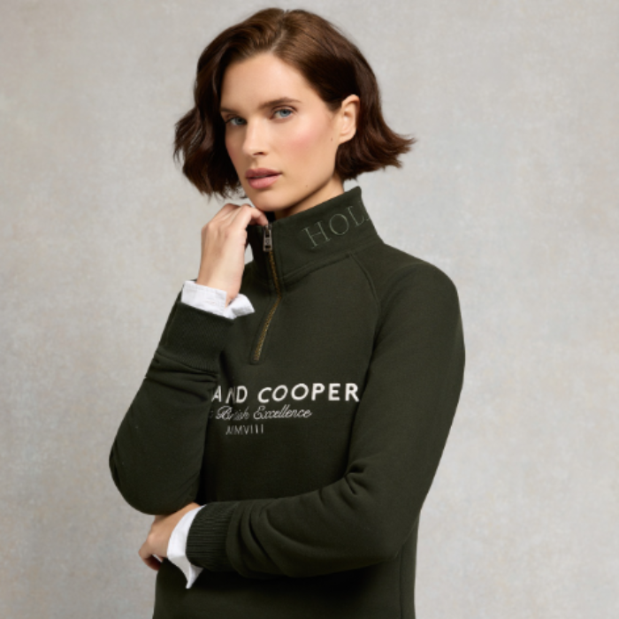 Holland Cooper Sporting Goods Zip Henley variant Racting Green. A woman wearing a dark olive green quarter-zip pullover sweater with white embroidered branding and cuffs, styled over a white collared shirt, against a neutral gray background.