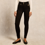 Holland Cooper Jodhpur Jean Variant Black. A model wearing black skinny jeans with gold button and logo details, paired with a white long-sleeve top and black flats, against a textured beige studio background.
