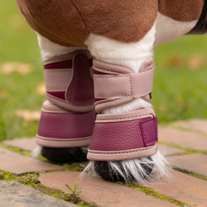 LeMieux AW26 Toy Pony Mesh Brushing Boots Variant Colour Blackberry. A close up of a toy pony legs wearing equestrian leg wraps in blackberry colour on faded garden background.