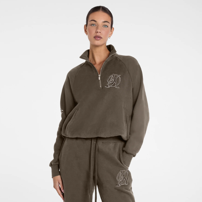 A model wearing a matching brown fleece tracksuit set, featuring a cropped half-zip pullover with a silver geometric logo on the chest and matching jogger pants with a drawstring waist and pocket.