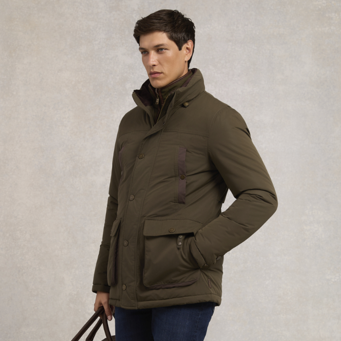 Holland Cooper Stamford Field Coat (Men's) Variant khaki. A man wearing an olive green puffer jacket with a high collar, front button closure, and a vertical chest pocket, standing against a textured light gray background.