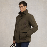 Holland Cooper Stamford Field Coat (Men's) Variant khaki. A man wearing an olive green puffer jacket with a high collar, front button closure, and a vertical chest pocket, standing against a textured light gray background.