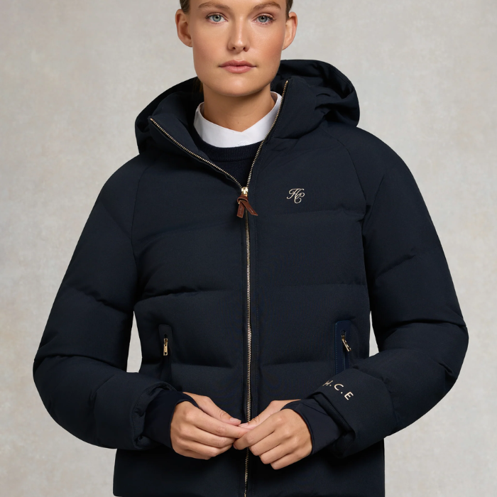 Holland Cooper Chiltern Yard Jacket Variant Ink Navy. A model wearing a dark navy hooded puffer jacket with gold-toned zippers and a small embroidered logo on the left chest, set against a neutral textured studio background.