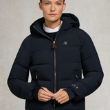 Holland Cooper Chiltern Yard Jacket Variant Ink Navy. A model wearing a dark navy hooded puffer jacket with gold-toned zippers and a small embroidered logo on the left chest, set against a neutral textured studio background.