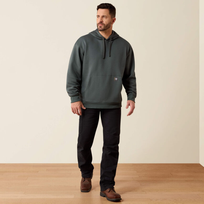 A man wearing a dark green pullover hoodie with black drawstrings, black pants, and brown boots, standing in a studio with a beige wall and light wood floor.