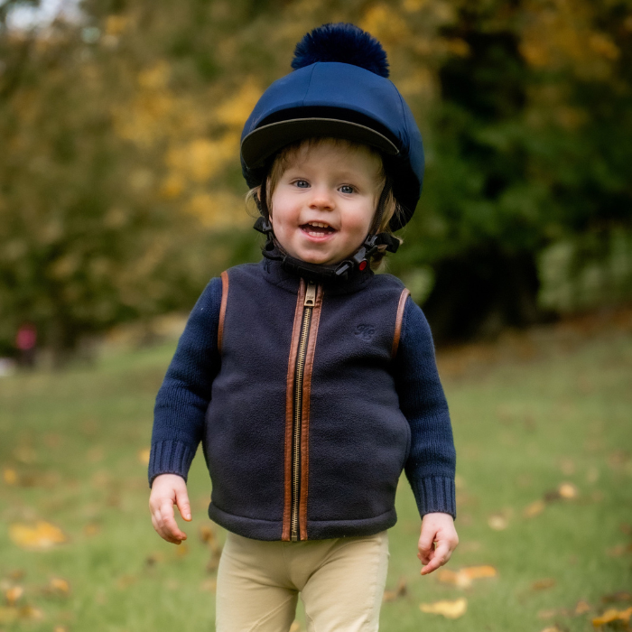 Holland Cooper Little Country Fleece Gilet Varient Ink Navy. Young boy wearing a blue fleece gilet with brown leather trim detailing and a small HC logo. The boy is wearing a navy jumper underneath and cream johdpurs in a woodland setting. 