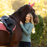 LeMieux AW26 Heidi Hooded Mid Layer Variant Colour Aloe.
A woman in a sage green long-sleeve athletic top and navy pants stands beside a dark brown horse outdoors. The horse is wearing pink ear muffs and a black bridle with a black saddle and pink-and-white saddle pad.